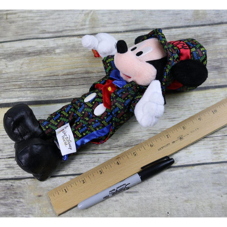 May include: A plush Mickey Mouse wearing a black tuxedo with a colorful 2000 pattern, a red bow tie, and a black top hat with a red 2000 band. The plush is holding a small red object in its right hand. The plush is labeled "Walt Disney World".