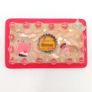 May include: A pink and gold mosaic tile art piece with a pink cupcake, a bottle cap with the word "Sweet" on it, and a butterfly.