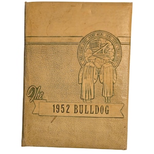 Puede incluir: Un anuario vintage con una cubierta texturizada de color marrón claro. La cubierta presenta un diseño en relieve con dos figuras con togas de graduación, una bandera estadounidense y el texto "1952 BULLDOG". También está presente la palabra "The".