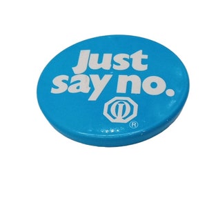 Puede incluir: Un botón redondo azul con el texto "Just say no." en letras blancas. El botón también tiene un círculo blanco con una letra "Q" dentro y un símbolo de marca registrada.