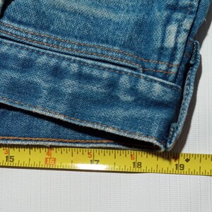 Puede incluir: Primer plano del borde inferior de una chaqueta vaquera azul con una cinta m&eacute;trica amarilla que muestra de 15 a 19 pulgadas.
