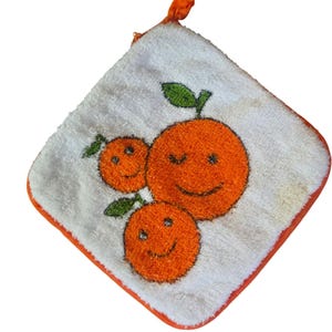 Op de afbeelding: Witte vierkante keukendoek met een oranje rand en een ophanglus. De handdoek heeft een lachend sinaasappelvruchtenontwerp met groene bladeren. De sinaasappels hebben zwarte lachende gezichten.