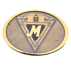 May include: Oval-shaped metal belt buckle with a gold-colored border. The buckle features a bronze-colored center with a black triangle containing a stylized gold letter "M". Above the triangle is a rectangle with the number "5".