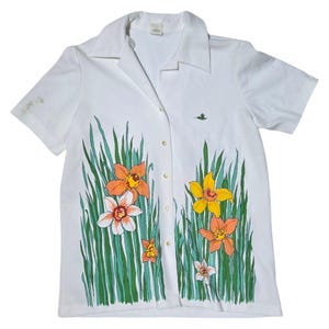 May include: White short-sleeved button-up shirt with a floral print. The print features orange, yellow, and white daffodils with green leaves. A small green hummingbird is printed in the upper right corner.