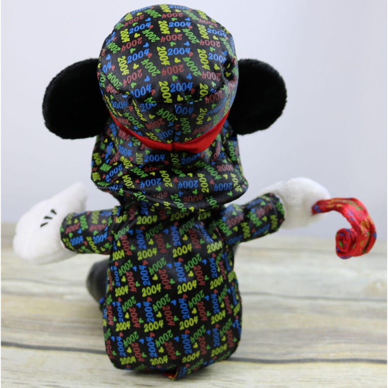 May include: A black plush Mickey Mouse wearing a black and red hat with a repeating pattern of "2004" in various colors. Mickey is holding a red and white curled party horn.