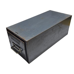 May include: A rectangular, gray metal file box with a handle and a slot on top. The box has a label on the front with text. The metal has a worn appearance.