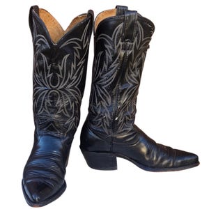 May include: A pair of black cowboy boots with intricate white stitching. The boots have pointed toes and stacked heels. The leather appears worn, adding to their vintage appeal. The boots are a classic Western wear item.