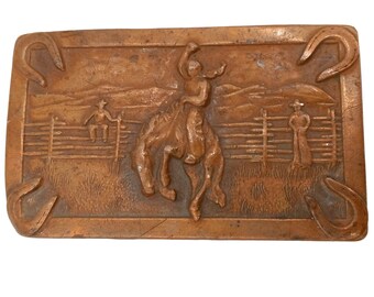 Puede incluir: Una hebilla de cinturón de color cobre con un diseño en relieve de un vaquero montando un bronco que se resiste. La hebilla está rodeada por dos herraduras.
