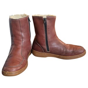 May include: A pair of brown leather boots with a zipper closure and a tan sole. The boots have a soft, light-colored lining visible at the top. The boots are designed for cold weather.