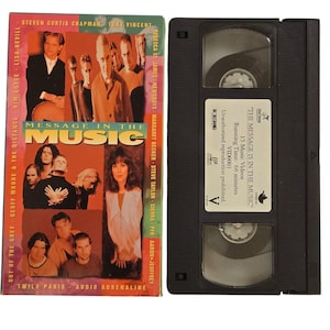 Puede incluir: Una caja de cinta VHS con fondo negro y una imagen colorida de un grupo de personas. El título del video es "Message in the Music" y presenta 15 videos musicales con una duración total de 66 minutos.