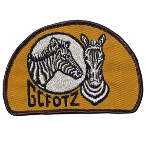 Könnte beinhalten: Gestickter Aufnäher mit zwei Zebra-Köpfen auf senfgelbem Hintergrund, mit braunem Rand. Die Zebras sind in einem kreisförmigen Design mit schwarz-weißen Streifen angeordnet. Der Text "GCFOTZ" ist in Schwarz gestickt.