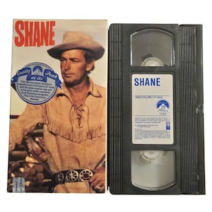 以下が含まれることがあります： 映画「シェーン」のヴィンテージVHSテープとボックスセット。ボックスにはカウボーイハットをかぶった男性と赤いタイトルが描かれています。VHSテープは黒色で、「SHANE」と書かれた白いラベルが付いています。