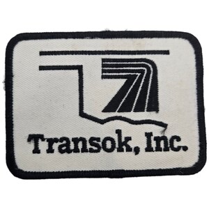 May include: A white embroidered patch with black stitching. The patch features a stylized graphic of a road with a curved section and the text "Transok, Inc."
