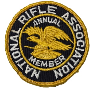 Pode incluir: Remendo circular com fundo preto e borda dourada. O remendo apresenta o texto "NATIONAL RIFLE ASSOCIATION ANNUAL MEMBER" em letras brancas. Um emblema de águia dourada está no centro.