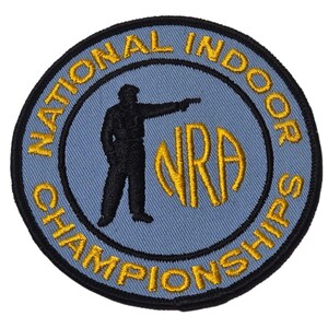 May include: A circular patch with a light blue centre, black border, and gold lettering. The text reads "NATIONAL INDOOR CHAMPIONSHIPS" and "NRA". A black silhouette of a person aiming a pistol is in the centre.