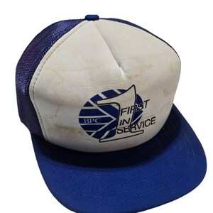 Puede incluir: Una gorra de camionero blanca y azul con un logotipo azul y blanco que dice "BPC First in Service". La gorra tiene una parte trasera de malla azul y una visera azul.