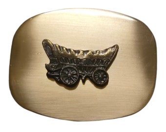 Könnte beinhalten: Eine goldfarbene Gürtelschnalle aus Metall mit einem erhabenen Design eines Planwagens. Der Wagen ist mit Rädern und einem Holzrahmen detailliert.
