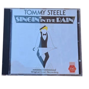 May include: CD case for the musical "Singin' in the Rain" by Tommy Steele. The cover features a cartoon illustration of a man in a tuxedo and bowler hat holding an umbrella, with the title in a stylized font. The words "Original Cast Recording" are also visible.