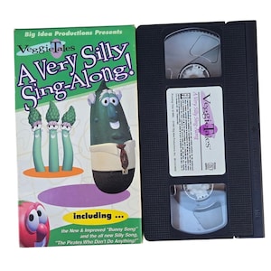 May include: A VHS tape and its cover for "VeggieTales: A Very Silly Sing-Along!" The cover is green, purple, and white, featuring cartoon vegetables. The VHS tape is black with a white label.