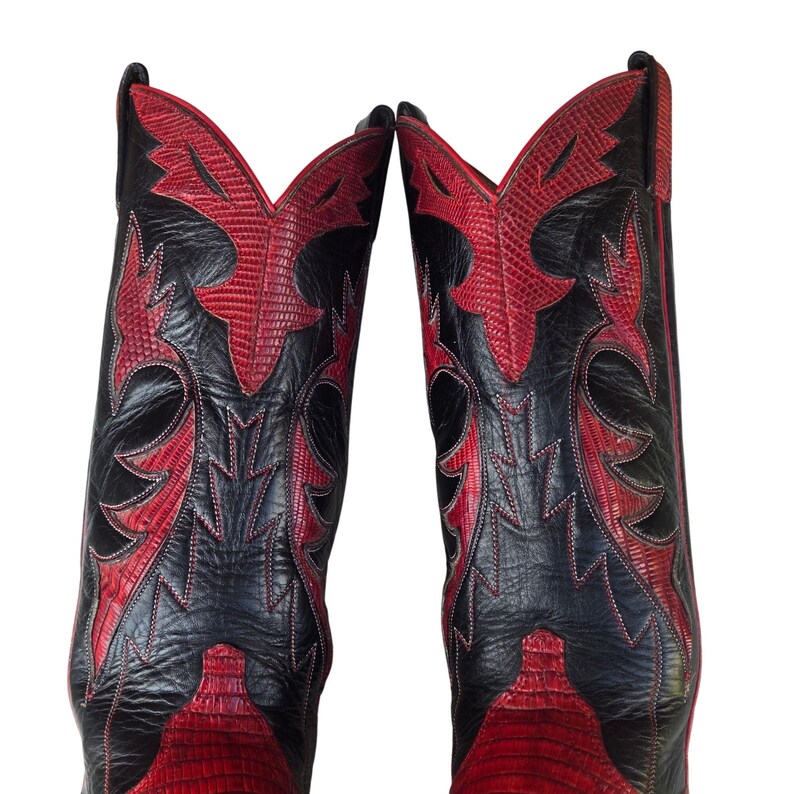 May include: A pair of cowboy boots in black and red. The boots feature a textured leather finish, with red panels and decorative stitching against a black background. The design is eye-catching.