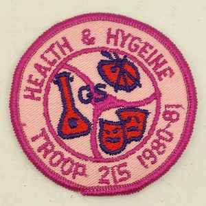 Può includere: Un cerotto rotondo rosa con un bordo rosa. Il cerotto ha uno sfondo bianco con il testo "Health & Hygiene Troop 215 1999-2000" in un cerchio rosa. All'interno del cerchio ci sono tre immagini ricamate in rosso e viola: una beuta, una farfalla e una maschera.