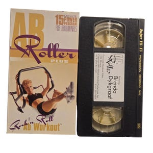 Puede incluir: Un conjunto vintage de cinta VHS y caja para el programa de ejercicios "AB Roller Plus". La caja es crema y dorada con una mujer usando el rodillo abdominal. La cinta VHS es negra con una etiqueta blanca.