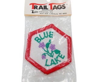 May include: A red hexagonal patch with white fabric and embroidered text that reads "BLUE LAKE" in green and purple thread. The patch has a small embroidered flower design with green leaves and purple flowers.