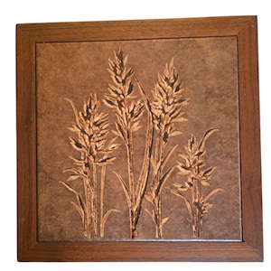 May include: Square decorative tile with a wheat stalk design. The tile is a mottled brown color, with the wheat stalks in a lighter brown. The tile is set in a dark brown wooden frame.