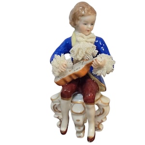 May include: A porcelain figurine of a seated figure playing a stringed instrument. The figure wears a blue jacket with white lace trim, burgundy breeches, and white stockings. The figurine is seated on a white and gold ornate chair.