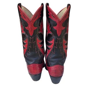 May include: Pair of black and red cowboy boots. The boots have a black leather upper with red decorative stitching and accents. The toe and heel are red, and the boots have a classic Western design. A stylish footwear choice.