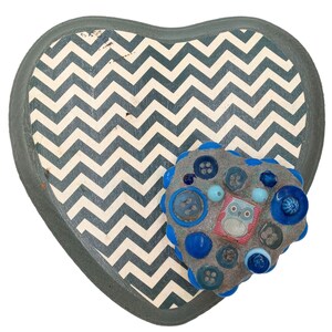 May include: A heart-shaped tray with a gray and white chevron pattern. A small heart-shaped mosaic is attached to the tray. The mosaic is made of blue and gray buttons, beads, and a small pink owl charm.