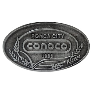 May include: Oval-shaped, silver-toned metal belt buckle. The buckle features the text "PONCA CITY" above the "CONOCO" logo, with the year "1983" below. Wheat stalks frame the logo.