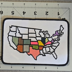 Könnte beinhalten: Ein rechteckiger Aufnäher mit einer Karte der Vereinigten Staaten. Die Staaten sind schwarz umrandet und mit verschiedenen Farben wie Rosa, Grün und Braun gefüllt. Der Aufnäher hat einen weißen Hintergrund und einen schwarzen Rand. Ca. 15 cm lang.