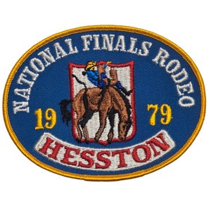 Peut inclure: Un écusson brodé ovale avec un fond bleu et une bordure dorée. Il présente un cheval brun au galop avec un cavalier en bleu et jaune, centré dans un écusson blanc. Le texte indique NATIONAL FINALS RODEO, 19 79 et HESSTON en rouge.