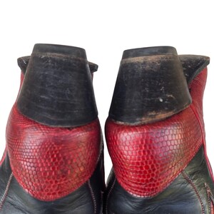 May include: Close-up of two cowboy boots. The boots have a black leather upper and a red, textured vamp. The heel is black and the stitching is visible. The boots are set against a white background.