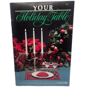Może przedstawiać: Antyczna książka zatytułowana "Your Holiday Table" z świątecznym obrazem nakrycia stołu z srebrnym świecznikiem z trzema zapalonymi białymi świecami, czerwonymi gwiazdami betlejemskimi, zielonym wieńcem z czerwonymi jabłkami i szyszkami oraz czerwoną serwetką złożoną w ozdobny kształt na białym talerzu.