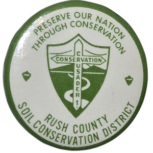 May include: A circular button in white and green, featuring the text "PRESERVE OUR NATION THROUGH CONSERVATION" around the top edge. The centre displays a green shield with the words "CONSERVATION" and "USADER". The bottom reads "RUSH COUNTY SOIL CONSERVATION DISTRICT".