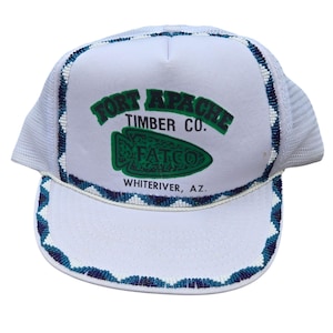May include: White trucker hat with a mesh back and a beaded brim. The front panel features the text "FORT APACHE TIMBER CO." in green, along with a green arrowhead logo and the text "FATCO". The text "WHITE RIVER, AZ." is printed below the logo.