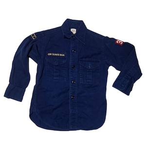 Op de afbeelding: Een vintage marineblauw Cub Scouts B.S.A. overhemd met knopen. Het overhemd heeft twee borstzakken, een kraag en lange mouwen. De tekst "CUB SCOUTS B.S.A." is op de borst geborduurd. Er zitten patches op de mouwen.