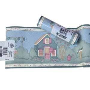 May include: A roll of wallpaper border featuring a quaint house with pink shutters and a blue roof, surrounded by green trees and pink hearts. The border has a light blue background with a repeating pattern of small hearts and arrows.