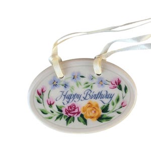May include: Cream-coloured oval ceramic ornament with the words "Happy Birthday" in script, encircled by a floral wreath of pink, yellow, and blue flowers. It is suspended by a white ribbon.