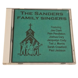 May include: A CD case featuring "The Sanders Family Singers." The cover is teal with a church illustration and the band's name in white. The CD case is clear plastic.