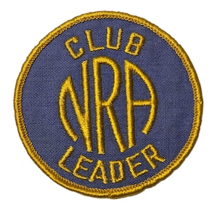 May include: A circular embroidered patch featuring a navy blue background and gold lettering and border. The text reads "CLUB NRA LEADER". The patch is likely for a club or organization, with a gold border.