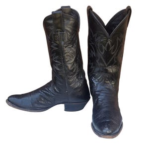 Puede incluir: Un par de botas de estilo western negras con un patrón texturizado de piel de lagarto en el pie y una caña lisa con costuras tonales intrincadas. Las botas tienen punta afilada, tacón apilado y tiradores en la parte superior de la caña.