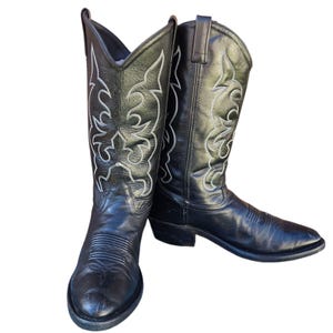 May include: A pair of black leather cowboy boots with decorative white stitching. The boots have pointed toes and stacked heels. The stitching forms a floral pattern on the boot shafts.