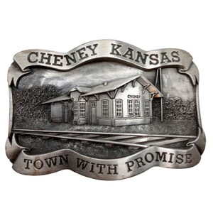 Könnte beinhalten: Eine silberne Gürtelschnalle mit einem Reliefbild eines Bahnhofsgebäudes und dem Text "Cheney Kansas Town With Promise".