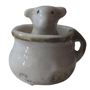 May include: A small white ceramic pot featuring a sculpted animal head emerging from the center. The pot has a tan rim, a side handle, and faint black text printed on the lower exterior surface. The animal figure has two small black dots for eyes.