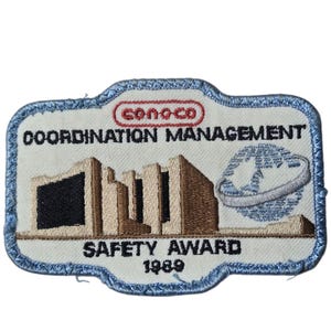 May include: Embroidered patch with a light blue border and a white background. The patch features the Conoco logo, the words "COORDINATION MANAGEMENT" and "SAFETY AWARD 1989". The design includes a stylized building and a globe.