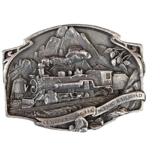 May include: A silver-coloured belt buckle with a detailed relief of a steam train, mountains, and the text "Cumbres & Toltec Scenic Railroad." The buckle has an ornate border and is likely made of metal.