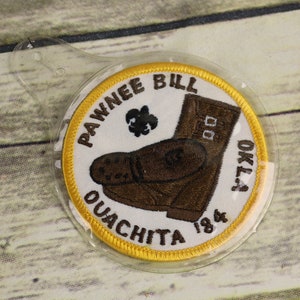 Może przedstawiać: Okrągła naszywka z napisem "Pawnee Bill Ouachita Okla 1984" i brązową ilustracją buta. Naszywka ma żółtą obwódkę i jest w przezroczystym plastiku.